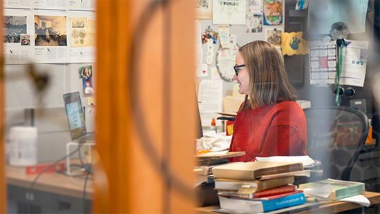 A library staff member works at a desktop computer in an office. Papers cover the wall. 