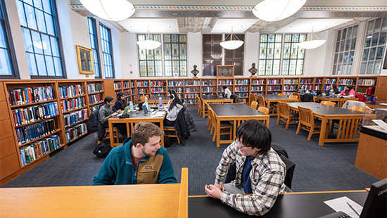 Two students talk at a desk in a library study space. Tables with students fill the room, and bookshelves line the outer walls.