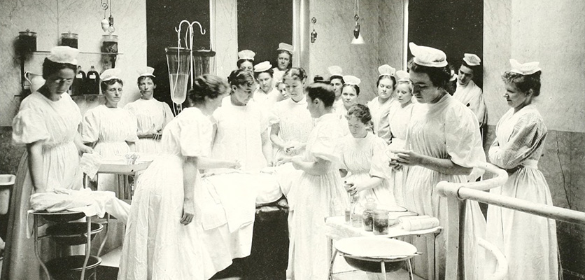 A group of women medical trainees, wearing dresses and hats, gather around a surgical demonstration at Marie Mergler's clinic at the Woman's Hospital.